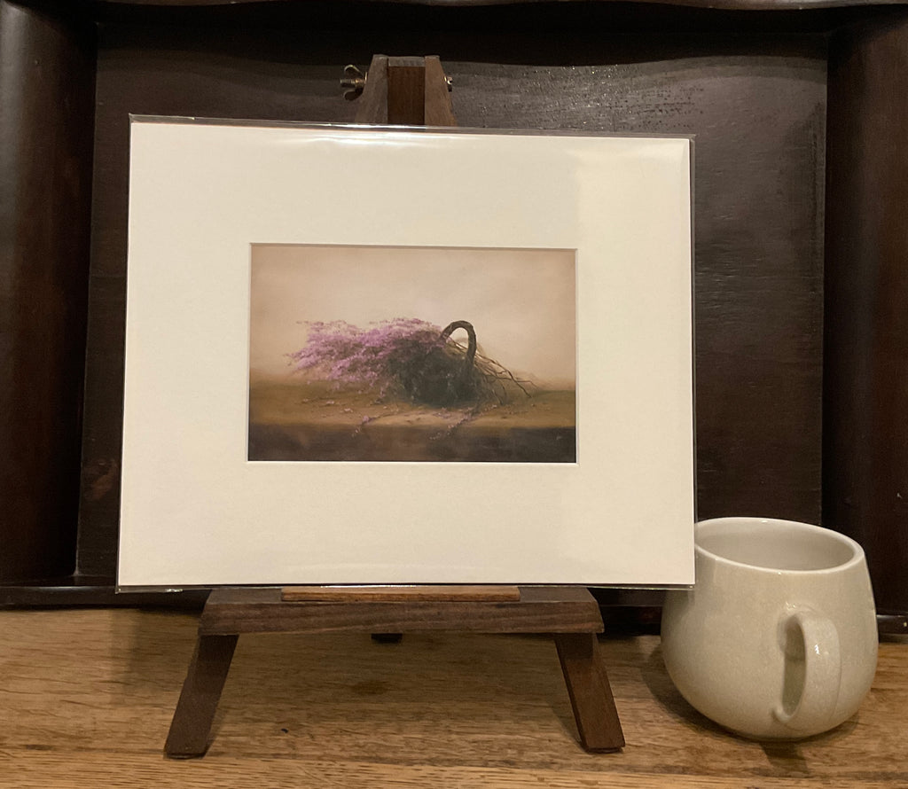 A photo of a Giclée print from Judith Levin original oil painting Heather Basket depicting heather in a beasket, on a mini easel next to a mug.