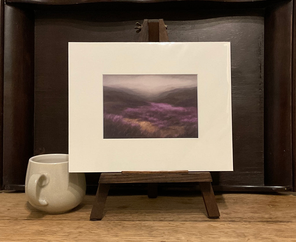 A photo of a Giclée print from Judith Levin original oil painting Heather Valley, depicting heather moorland, on a mini easel next to a mug.
